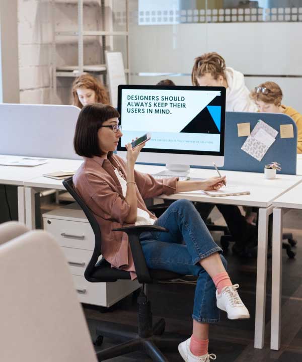 A woman sitting in an office chair, holding a smartphone to her mouth, appears to be speaking into it. She is surrounded by desks and computers, with a large monitor in the background displaying the text "Designers should always keep their users in mind." Several other people are working at their desks in the background.